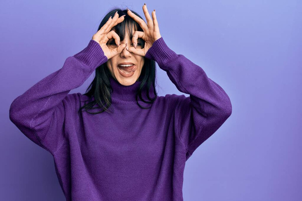 Young brunette woman with bangs wearing turtleneck sweater doing ok gesture like binoculars sticking tongue out, eyes looking through fingers. crazy expression.  - Фото, изображение
