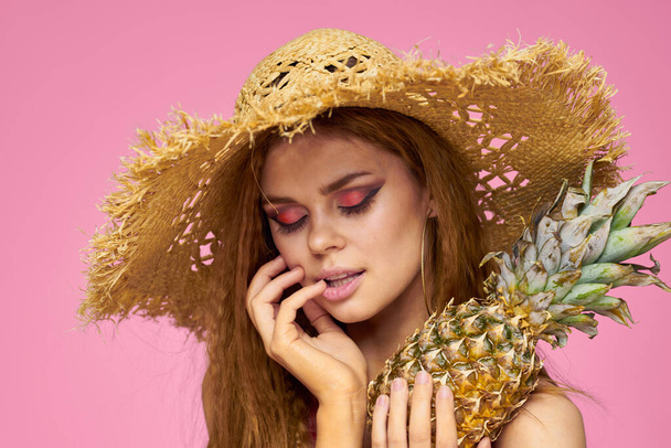 Woman with a gun in hands of a straw hat bright makeup exotic fruits summer pink background - Foto, imagen