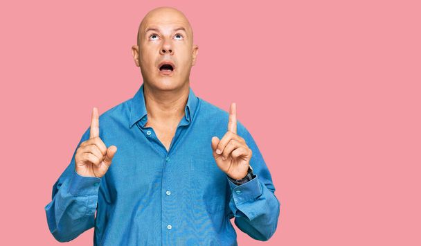 Middle age bald man wearing casual clothes amazed and surprised looking up and pointing with fingers and raised arms.  - Photo, Image