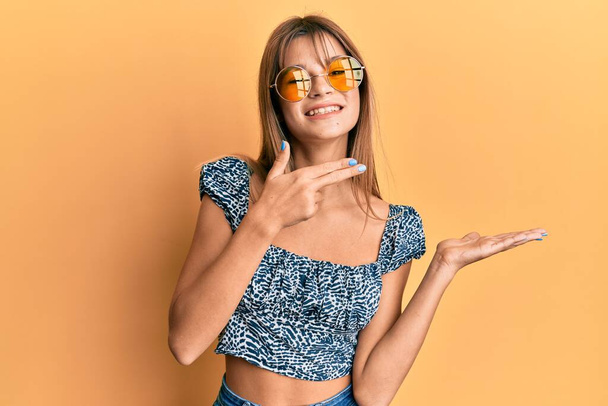 Teenager běloška dívka nosí módní žluté sluneční brýle ohromen a usmívá se do kamery při prezentaci s rukou a ukazuje prstem.  - Fotografie, Obrázek