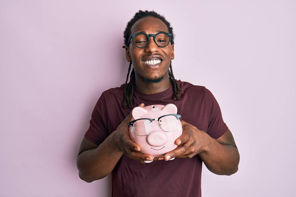 African american man with braids holding piggy bank with glasses celebrating crazy and amazed for success with open eyes screaming excited.  - Photo, Image