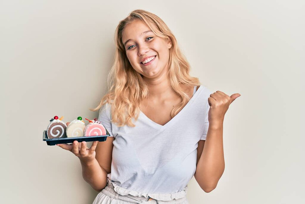Young blonde girl holding cake sweets pointing thumb up to the side smiling happy with open mouth  - Photo, image