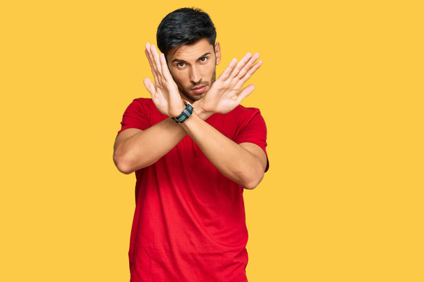 Fiatal jóképű férfi visel alkalmi piros póló elutasítás kifejezés keresztbe karok csinál negatív jel, dühös arc  - Fotó, kép
