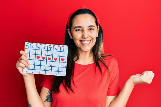 Mujer hispana joven sosteniendo el calendario del corazón gritando orgullosa, celebrando la victoria y el éxito muy emocionada con el brazo levantado  - Foto, Imagen
