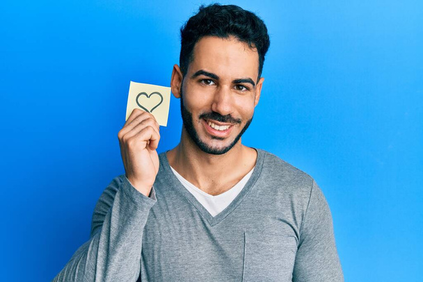 Νεαρός Ισπανόφωνος κρατώντας μια υπενθύμιση καρδιάς που δείχνει θετικός και χαρούμενος στέκεται και χαμογελά με ένα σίγουρο χαμόγελο που δείχνει τα δόντια  - Φωτογραφία, εικόνα