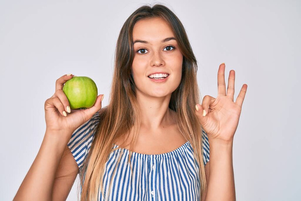 Mooie blanke vrouw met groene appel doet ok teken met de vingers, glimlachend vriendelijk gebaar uitstekend symbool  - Foto, afbeelding