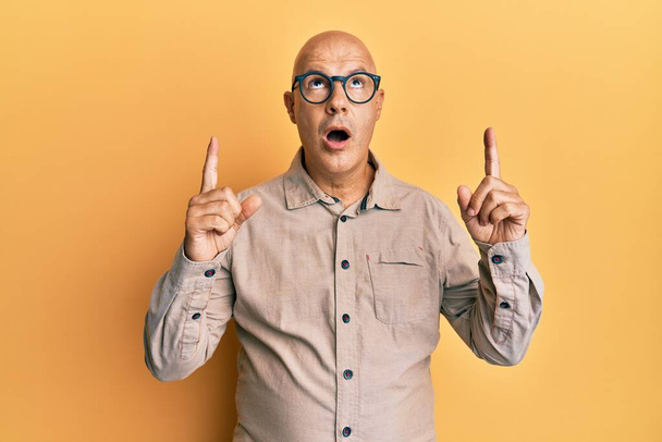Mann mittleren Alters mit Glatze, lässiger Kleidung und Brille staunt und überrascht und zeigt mit erhobenen Armen und Fingern nach oben.  - Foto, Bild