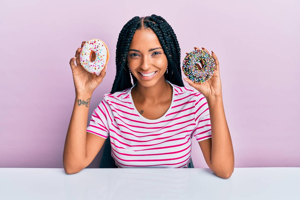 Schöne hispanische Frau hält leckere bunte Donuts lächelnd mit einem fröhlichen und kühlen Lächeln im Gesicht. Zähne zeigen.  - Foto, Bild