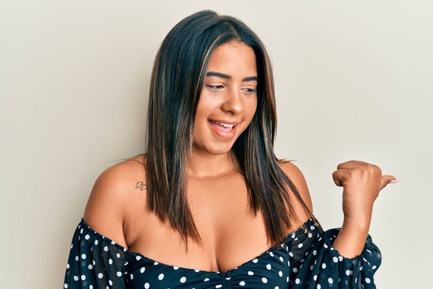 Giovane ragazza latina indossa abiti casual puntando il pollice verso l'alto per il lato sorridente felice con la bocca aperta  - Foto, immagini
