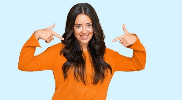 Красивая брюнетка молодая женщина в обычном оранжевом свитере выглядит уверенно с улыбкой на лице, указывая на себя пальцами гордой и счастливой.  - Фото, изображение
