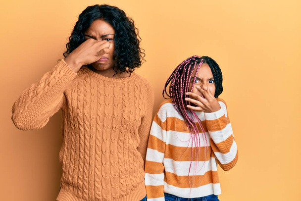Beautiful african american mother and daughter wearing wool winter sweater smelling something stinky and disgusting, intolerable smell, holding breath with fingers on nose. bad smell  - Fotó, kép