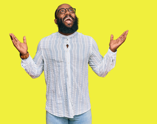 Νεαρός Αφροαμερικάνος που φοράει επιχειρηματικό πουκάμισο και γυαλιά γιορτάζει τρελός και τρελός για την επιτυχία με τα χέρια σηκωμένα και κλειστά μάτια ουρλιάζοντας ενθουσιασμένος. έννοια νικητή  - Φωτογραφία, εικόνα