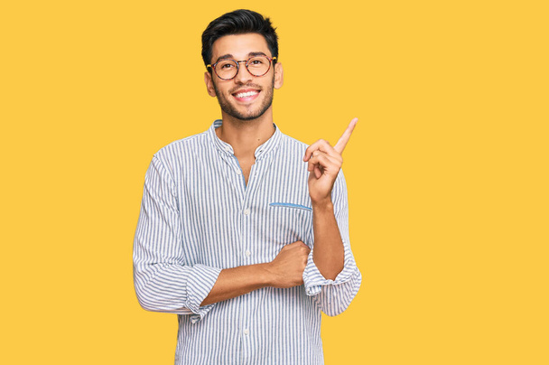 Young handsome man wearing casual clothes and glasses with a big smile on face, pointing with hand and finger to the side looking at the camera.  - Photo, Image