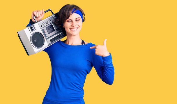 Gyönyörű fiatal nő rövid haj visel edzőruha és tartja boombox mutató ujját, hogy egy önmosolygó boldog és büszke  - Fotó, kép