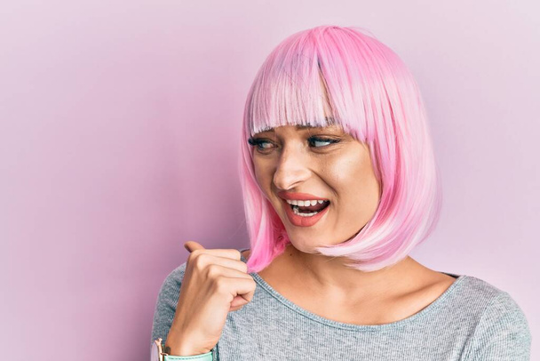 Joven mujer caucásica vistiendo peluca rosa sonriendo con la cara feliz mirando y señalando a un lado con el pulgar hacia arriba.  - Foto, Imagen