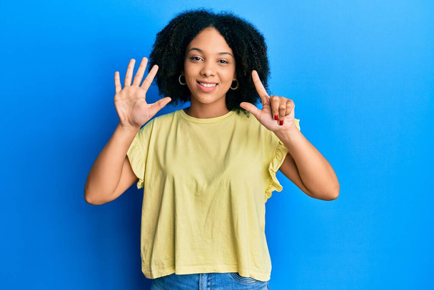 Junge afrikanisch-amerikanische Mädchen in lässiger Kleidung zeigt und zeigt mit den Fingern Nummer sieben, während sie selbstbewusst und glücklich lächelt.  - Foto, Bild