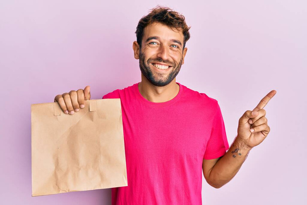 bel homme avec barbe tenant emporter sac en papier souriant heureux pointant avec la main et le doigt sur le côté  - Photo, image