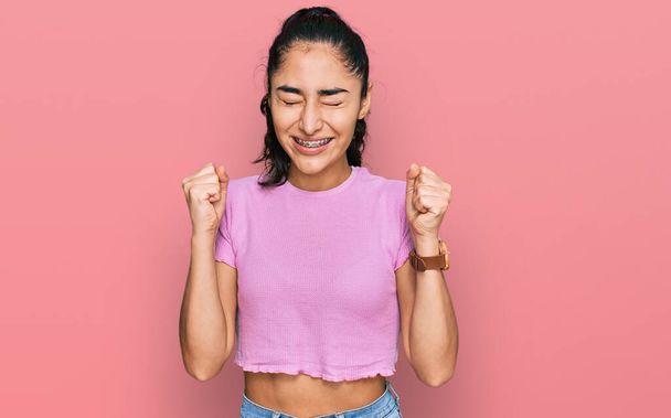 Adolescente hispanique avec appareil dentaire portant des vêtements décontractés excités pour le succès avec les bras levés et les yeux fermés célébrant la victoire en souriant. concept gagnant.  - Photo, image