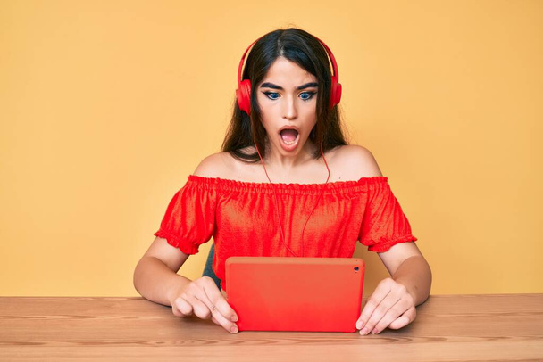 Brunette teenager girl using touchpad wearing headphones afraid and shocked with surprise and amazed expression, fear and excited face.  - Photo, Image