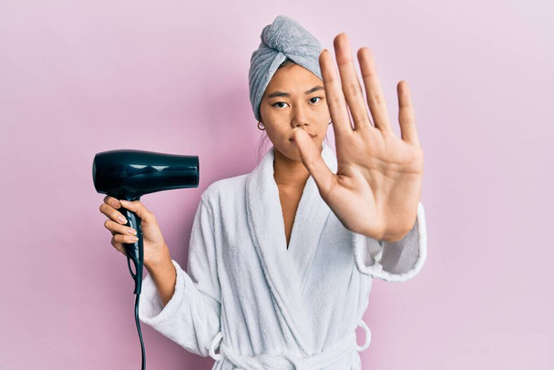 Young chinese woman wearing shower towel cap and bathrobe holding dryer with open hand doing stop sign with serious and confident expression, defense gesture  - Foto, afbeelding