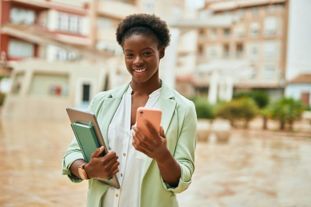 Νεαρή Αφροαμερικανή επιχειρηματίας χαμογελώντας ευτυχισμένη χρησιμοποιώντας smartphone στην πόλη. - Φωτογραφία, εικόνα