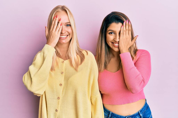 Δύο φίλοι στέκεται μαζί πάνω από ροζ φόντο καλύπτει το ένα μάτι με το χέρι, αυτοπεποίθηση χαμόγελο στο πρόσωπο και συγκίνηση έκπληξη.  - Φωτογραφία, εικόνα