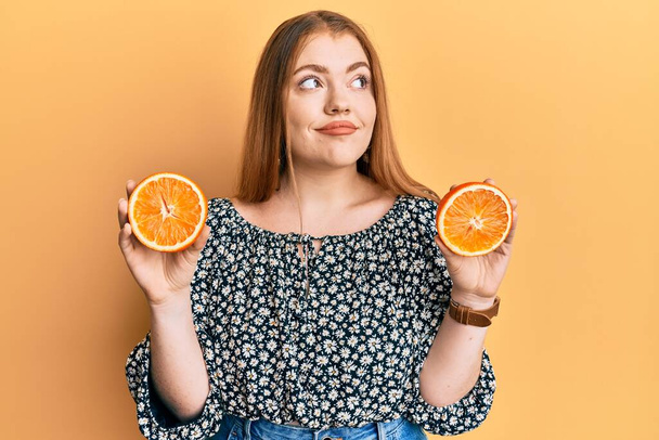 Jeune belle rousse femme tenant des tranches d'orange fraîche souriant regardant vers le côté et regardant loin en pensant.  - Photo, image
