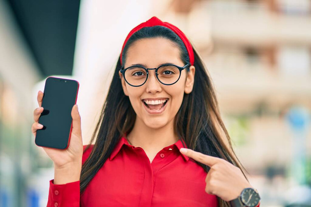 Mladá hispánka se šťastně usmívá ukazováním a ukazováním prstem na obrazovku smartphonu ve městě. - Fotografie, Obrázek