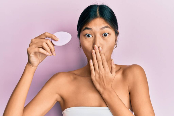 Fiatal kínai nő visel zuhany törölköző segítségével kozmetikai szivacs borító száj kézzel, sokkolt és fél a hiba. meglepett kifejezés  - Fotó, kép
