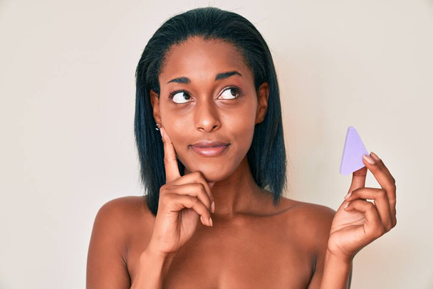 Молодая африканская американка, держащая губку для макияжа, всерьез думает о вопросе с рукой на подбородке, вдумчивая о запутанной идее  - Фото, изображение