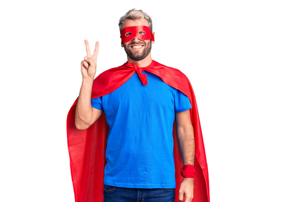 Junger blonder Mann trägt Superheldenmuster und zeigt mit Finger Nummer zwei nach oben, während er selbstbewusst und glücklich lächelt.  - Foto, Bild