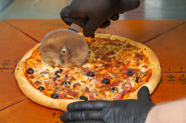 Cutting pizza with a round cutter knife. Close-up of delicious pizza being cut into pieces. - Photo, Image
