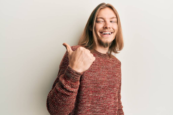 Hombre caucásico guapo con el pelo largo usando suéter de invierno casual apuntando pulgar hacia el lado sonriendo feliz con la boca abierta  - Foto, imagen