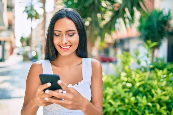 Молода латинська дівчина посміхається щаслива, використовуючи смартфон на вулиці міста
. - Фото, зображення