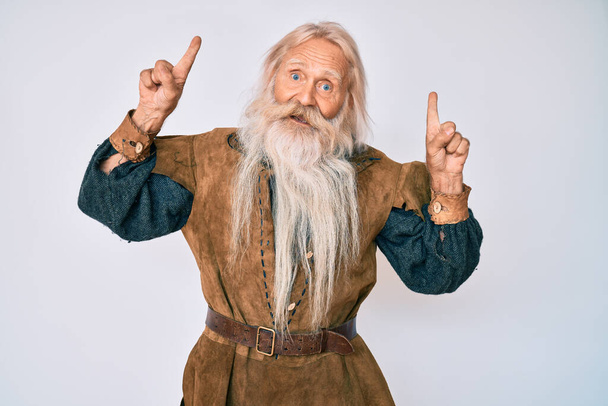 Idős öregember ősz hajjal és hosszú szakállal, viking tradicionális jelmezt visel, csodálkozva és meglepve, ujjal mutogatva és felemelt karral..  - Fotó, kép