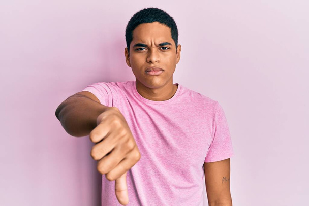 Νεαρός όμορφος Ισπανός με ροζ μπλουζάκι που δείχνει δυστυχισμένος και θυμωμένος δείχνοντας απόρριψη και αρνητικός με τους αντίχειρες κάτω. κακή έκφραση.  - Φωτογραφία, εικόνα