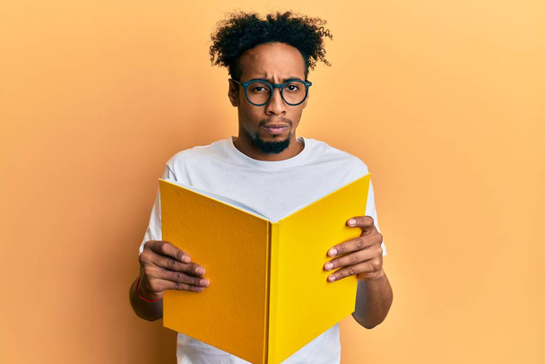 Joven afroamericano con barba leyendo un libro con gafas escéptico y nervioso, frunciendo el ceño molesto debido a un problema. persona negativa.  - Foto, imagen