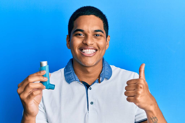 Jeune homme hispanique beau tenant inhalateur d'asthme médical souriant heureux et positif, pouce levé faire excellent et signe d'approbation  - Photo, image