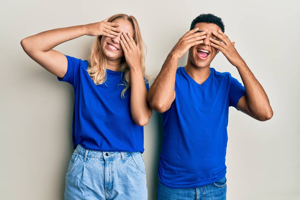 Jeune couple interracial portant des vêtements décontractés couvrant les yeux avec les mains souriantes gaies et drôles. concept aveugle.  - Photo, image
