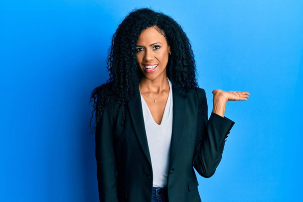 Middle age african american woman wearing business clothes smiling cheerful presenting and pointing with palm of hand looking at the camera.  - Photo, Image