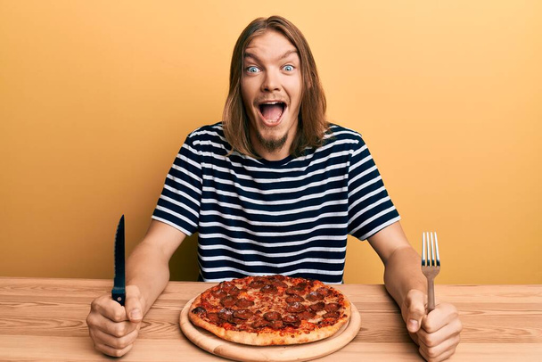 Jóképű fehér férfi hosszú hajjal eszik ízletes pepperoni pizza ünnepli őrült és csodálkozott a siker nyitott szemmel sikoltozó izgatott.  - Fotó, kép