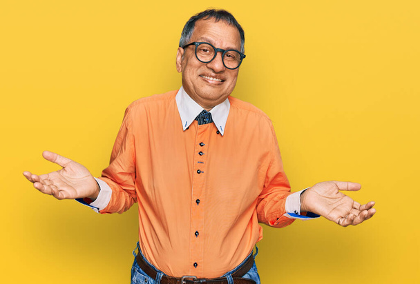Middle age indian man wearing casual clothes and glasses clueless and confused expression with arms and hands raised. doubt concept.  - Photo, Image