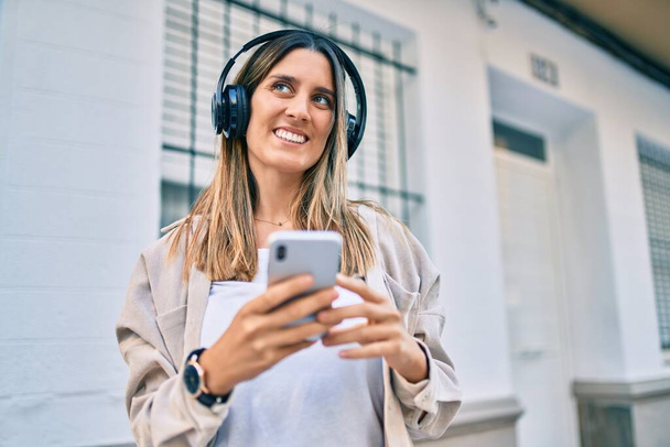 Joven mujer caucásica sonriendo feliz usando teléfono inteligente y auriculares en la ciudad. - Foto, imagen