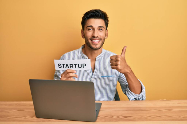 Junger gutaussehender Mann hält Startup-Nachricht als Marketing-Entwicklung lächelnd glücklich und positiv, Daumen hoch dabei hervorragende und Zustimmung Zeichen  - Foto, Bild