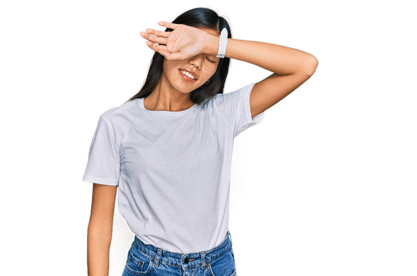 Beautiful young asian woman wearing casual white t shirt covering eyes with arm smiling cheerful and funny. blind concept.  - Foto, afbeelding