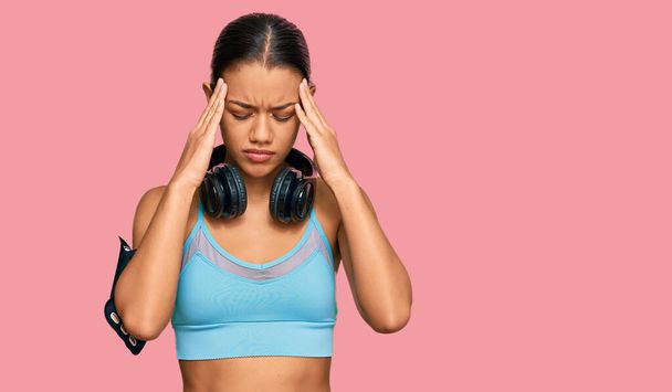 Schöne hispanische Frau in Sportbekleidung und Kopfhörer leiden unter Kopfschmerzen verzweifelt und gestresst, weil Schmerzen und Migräne. Hände auf den Kopf.  - Foto, Bild