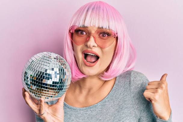 Jovem caucasiana vestindo peruca rosa segurando bola disco apontando polegar até o lado sorrindo feliz com a boca aberta  - Foto, Imagem