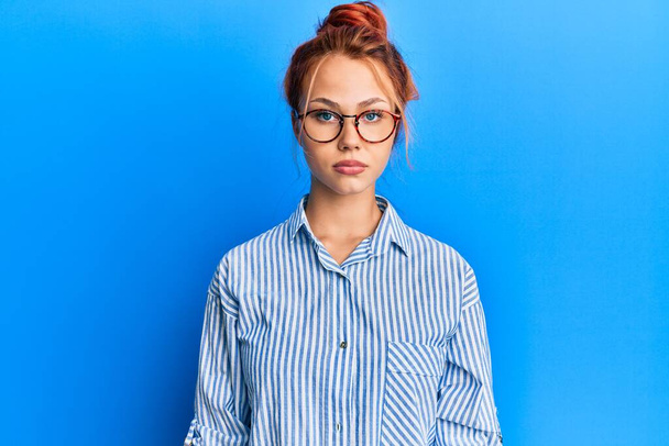 Young beautiful redhead woman wearing casual clothes and glasses over blue background with serious expression on face. simple and natural looking at the camera.  - Photo, Image