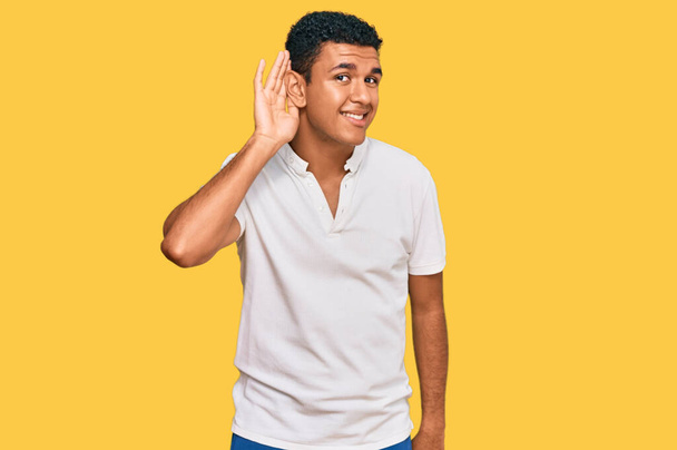 Jeune homme arabe portant des vêtements décontractés souriant avec la main sur l'oreille en écoutant une rumeur ou des commérages. concept de surdité.  - Photo, image