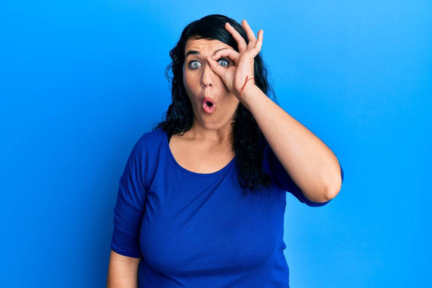 Plus grootte brunette vrouw draagt casual blauw shirt doet ok gebaar geschokt met verrast gezicht, oog kijken door vingers. ongelovige uitdrukking.  - Foto, afbeelding